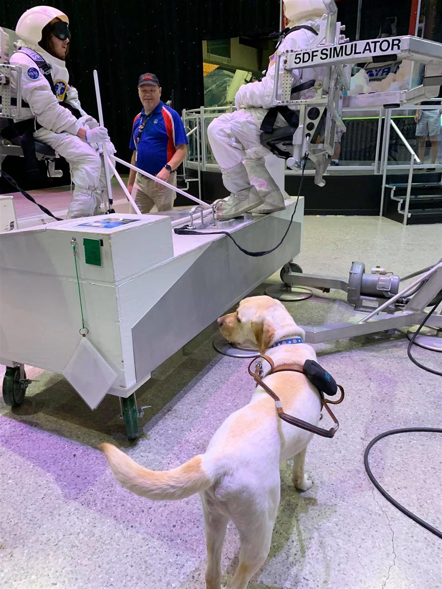 A guide dog looks on as two A.F.C.B. students sit on a space simulator. A third man stands between them.